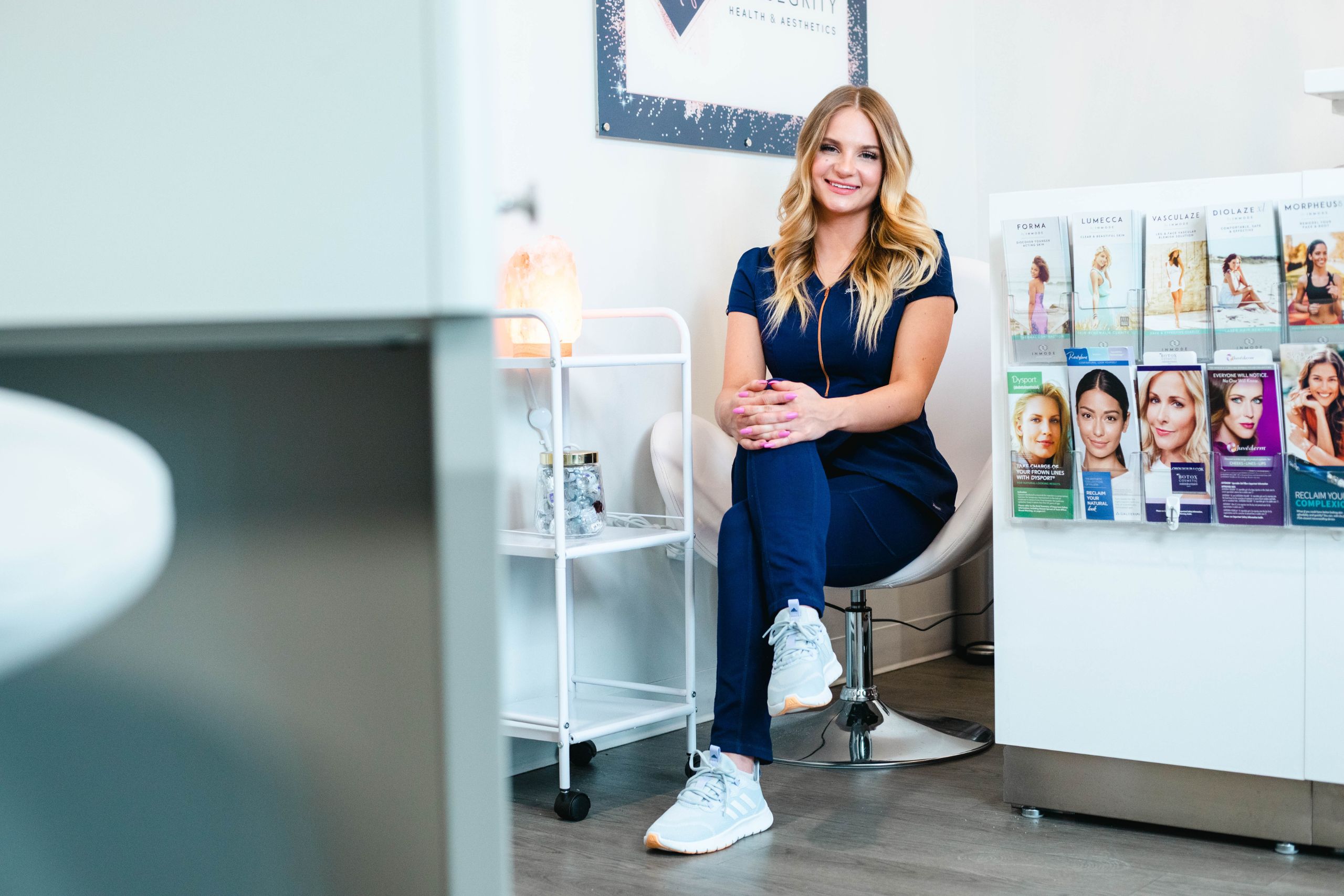 A medspa worker smiles at the camera while sitting in an office chair and crossing her legs.