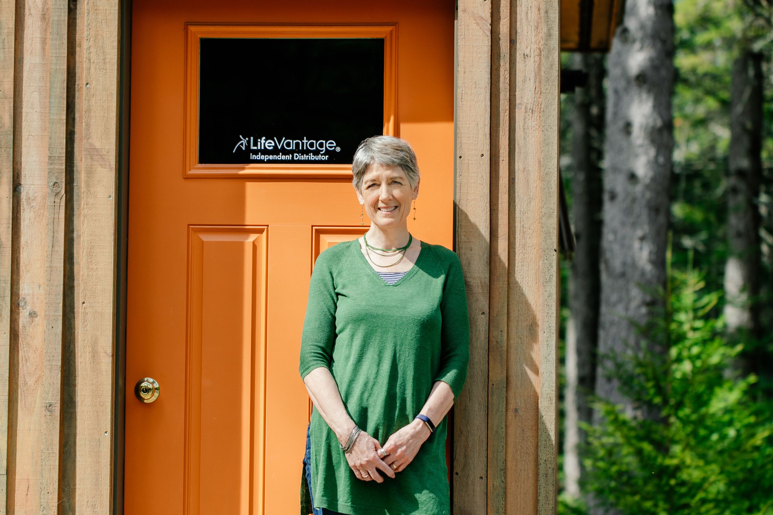 A woman poses outside her business in a green shirt. The building door reads, "LifeVantage: Independent Distributor."