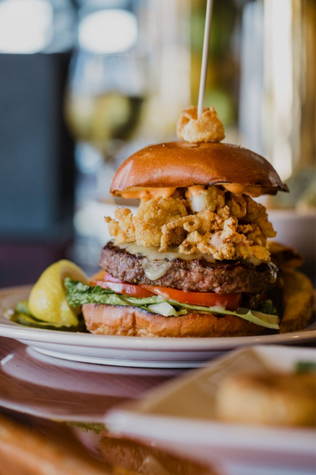A cheeseburger sits on a plate. It has fried clams, cheese, meat, tomatoes, and lettuce on it.