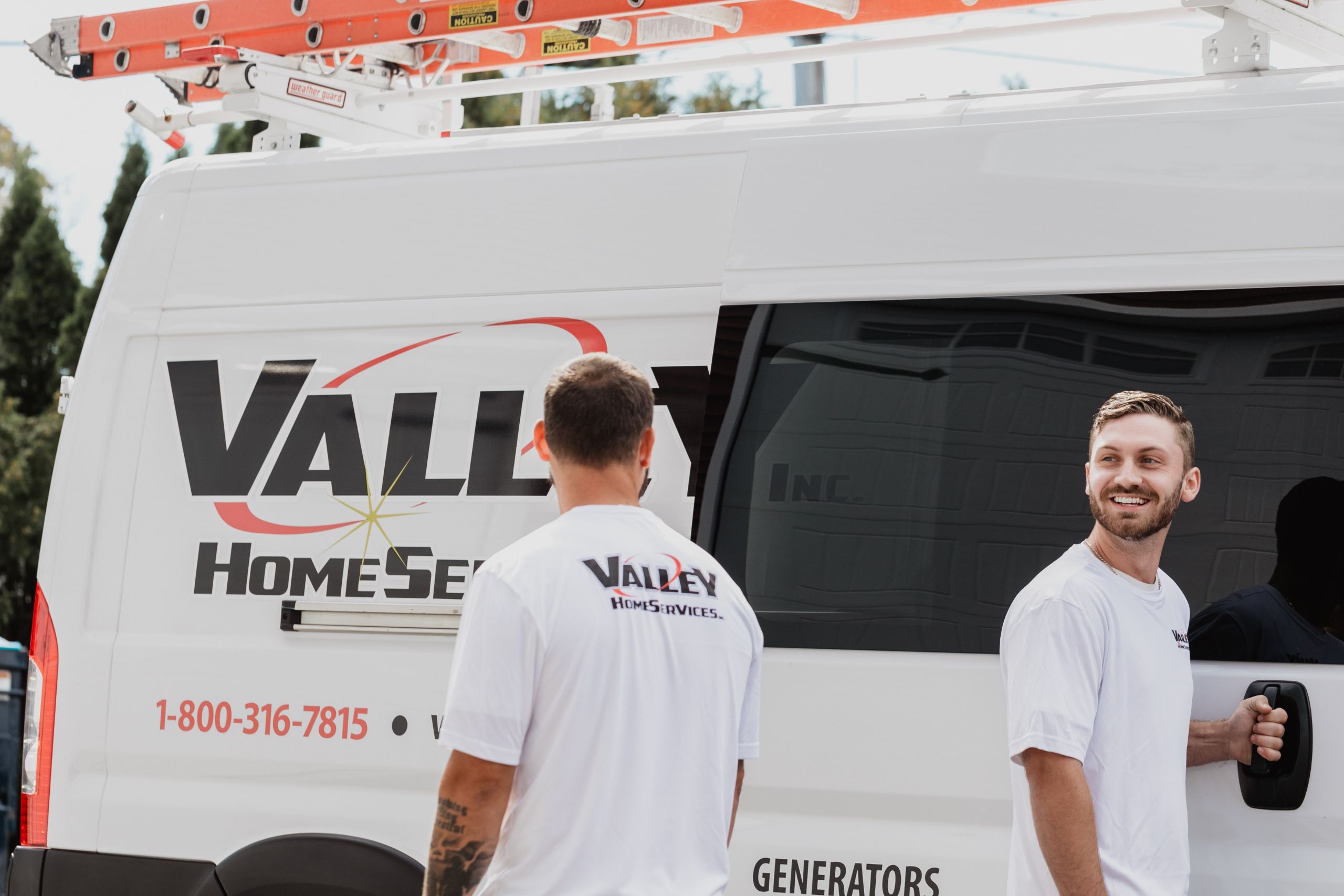 Two workers from Valley Home Services stand in front of their van. They smile at eachother.